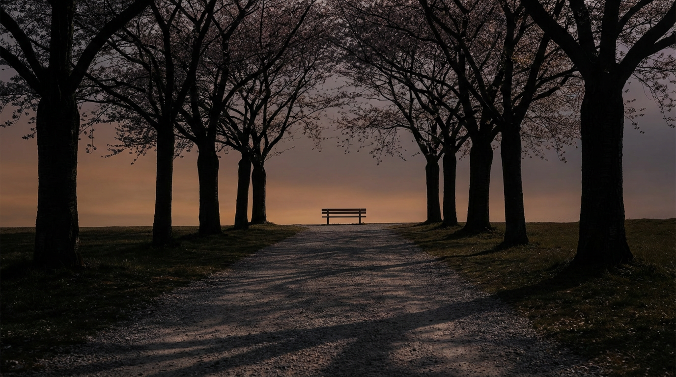 夕暮れの散歩道、並木の影が長く伸びている、道の先にベンチがひとつ見える、空はオレンジとグレーのグラデーション、人物は写らない